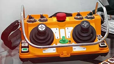 Close-up of an orange industrial remote control unit with two black joysticks, a red emergency stop button, and several toggle switches.